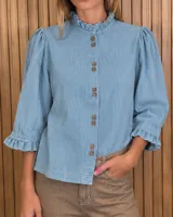 Camisa de denim celeste con cuello alto con volados, mangas tres cuartos abullonadas con puños con volados, y cierre frontal de botones.