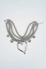 Collar multicapa compuesto por tres hileras de esferas metálicas plateadas. Presenta un dije central grande en forma de corazón irregular, acompañado por dijes más pequeños de corazones y monedas grabadas distribuidos a lo largo de las cadenas.