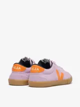 Championes Veja modelo Volley, confeccionados en gamuza color lila con detalles en naranja vibrante en el logo lateral y talón. Presentan suela de goma color caramelo y cierre con cordones al tono.