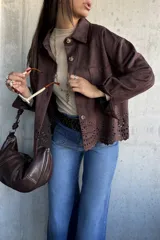 Chaqueta de gamuza color chocolate, con corte recto, cuello clásico y cierre frontal con botones. Presenta un diseño calado decorativo en el ruedo y bolsillos frontales tipo parche.