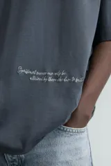 Camiseta de algodón color gris oscuro, corte relajado (relaxed fit), con cuello redondo y manga corta. Presenta dos inscripciones bordadas en hilo blanco en el frente.