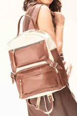 Mochila de cuero vacuno con diseño bicolor en tonos offwhite y tabaco texturado. Presenta un formato estructurado con dos bolsillos frontales con cierre metálico, correas laterales decorativas con hebillas y asa superior reforzada. El interior cuenta con forro de tela y compartimentos organizadores.