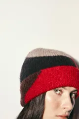 Gorro tipo beanie de punto con diseño de bloques de color en tonos rojo, negro y beige. Presenta un acabado con vuelta y textura suave.