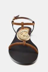 Sandalia plana con tiras de color marrón y un gran adorno metálico dorado circular en la parte frontal. Cuenta con cierre de hebilla ajustable al tobillo y suela plana de color negro.