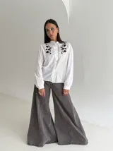 Camisa blanca de manga larga con cuello mao, cierre frontal de botones y detalle de volado. Presenta bordados florales en color marrón chocolate en la parte superior delantera.