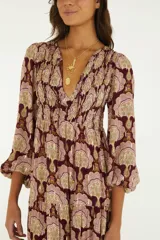 Vestido largo de bordó con estampado ornamental en tonos rosados y dorados, mangas tres cuartos abullonadas con puños elásticos, escote en V con cordones y falda escalonada con vuelo.