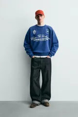 Sudadera de corte boxy fit en color azul con efecto lavado, cuello redondo y manga larga. Presenta estampado frontal con tipografía variada y gráficos estilo universitario. Interior afelpado y acabados en rib.
