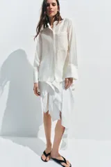Camisa de cuello mao color beige, confeccionada en tejido satinado con brillo sutil, manga larga con puños anchos y un bolsillo de parche en el pecho con detalles bordados tonales. Cierre frontal con botones ocultos bajo solapa.