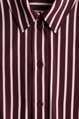 Camisa de manga larga con cuello clásico, cierre frontal abotonado y puños con botones. Presenta un diseño de rayas verticales en tonos bordó y crema, con bajo redondeado.