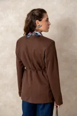 Chaquetón color beige de corte recto y largo, con cierre frontal mediante botones y lazo ajustable en la cintura. Presenta cuello en V, bolsillos laterales con solapa y mangas largas.