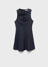 Vestido corto sin mangas con efecto denim, cuello redondo y detalle de lazos frontales. Presenta un diseño con tablas y cierre de cremallera en la parte posterior.