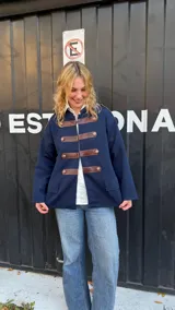 Campera de estilo militar en color azul marino, confeccionada en tejido de punto grueso. Presenta un diseño de cuello redondo, cierre frontal con tiras de cuero marrón y botones metálicos decorativos. Posee un corte recto y holgado.