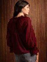 Blusa de plumeti color bordó, con cuello alto ajustable mediante lazo, mangas largas con puños elásticos y detalle de volados en las terminaciones.