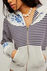 Buzo con capucha y cierre frontal de cremallera, confeccionado en tejido de punto rayado en tonos gris y azul, combinado con paneles lisos y detalles de estampado floral en la capucha y los puños. Presenta bolsillos frontales estilo canguro y diseño de largo corto.