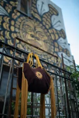 Bolso tipo tote de gamuza color negro con flecos y bordes en color salmón. Presenta un estampado de un sol con las iniciales de la marca en el centro. Tiene correa larga del mismo material.