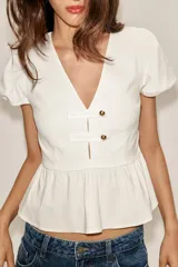 Top de color blanco con escote en pico, mangas cortas abullonadas y bajo con diseño peplum. Presenta detalle de botones dorados en la parte frontal y cierre lateral con cremallera oculta.