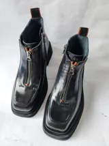 Bota corta de cuero negro con diseño de punta cuadrada y suela track. Presenta un cierre frontal central, detalles de pespuntes y una textura combinada en los laterales.