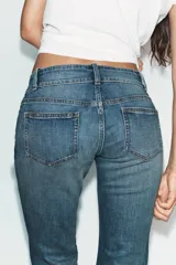 Jeans de tiro bajo con corte bootcut, confeccionados en denim con efecto lavado. Presentan costuras verticales marcadas en el frente, cierre frontal con cremallera, gancho metálico y detalle de trabilla con botones en la cintura.