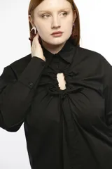 Camisa oversize de manga larga en color negro, confeccionada en algodón. Presenta cuello camisero clásico, puños abotonados y una abertura central ovalada con sistema de fruncido ajustable mediante botones y jaretas.