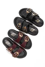 Sandalias planas tipo 'slide' de cuero con dos tiras anchas ajustables mediante hebillas doradas. La plantilla es negra con el logo 'MACU' en dorado. Se muestran en color negro y color bordó (vino tinto) charol.