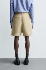 Bermuda color beige confeccionada en tejido de algodón y lino, con corte relaxed fit, cintura ajustable con cinturón de la misma tela, pliegues frontales, bolsillos delanteros y traseros, cierre frontal con cremallera y botón.