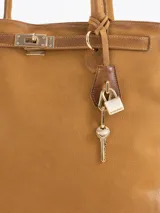 Bolso tipo tote color gris, confeccionado en cuero vacuno, con asas largas para hombro, detalles metálicos personalizados, cierre central y llavero con candado y llave colgante.