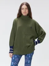Pullover de cuello tortuga en color verde oliva, con diseño holgado y mangas largas. Presenta puños acanalados en contraste azul marino con líneas horizontales claras y logo bordado en el pecho.