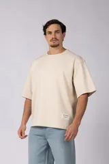 Remera de corte oversize color beige, con cuello redondo y etiqueta decorativa pequeña en el ruedo inferior.