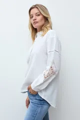Camisa blanca de corte recto con cuello clásico y cierre frontal de botones. Presenta mangas largas con detalles de encaje calado en los antebrazos y puños abotonados.