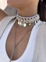 Gargantilla choker con hilo trenzado color blanco, adornada con conchas marinas tipo buzios y medallitas bañadas en oro y acero quirúrgico. Se cierra con cadena de acero quirúrgico y ganchito para ajustar el largo.