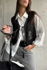 Chaleco de gamuza negra con bordados decorativos en color blanco y cierre frontal mediante cordones con borlas. Presenta un diseño corto y estilo boho.