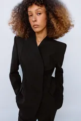 Blazer entallada de color negro con escote en pico, manga larga con hombreras y cierre frontal con botones ocultos. Presenta bolsillos con solapa en el delantero y detalle de nervios marcados en el tejido.