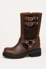 Bota de caña media confeccionada en una combinación de cuero y gamuza color marrón. Presenta un diseño estilo biker con dos correas ajustables con hebillas metálicas en la parte superior de la caña y una correa decorativa con argolla metálica y remaches en el tobillo. Cuenta con suela de goma dentada y taco cuadrado.