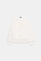 Blusa de georgette en color blanco, con escote en pico, detalle de volantes y lazada frontal. Presenta mangas largas abullonadas con puños ajustados y un bajo asimétrico con diseño tipo globo.