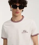 Remera blanca de algodón, corte baby tee, con cuello redondo y mangas cortas con ribete doble en bordó. Estampa pequeña con texto "The Staff" en el pecho.