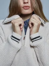 Sweater de tejido grueso color beige, con cuello alto y cierre frontal parcial. Presenta detalles de rayas negras en los puños y en el borde del cuello, con un diseño de estilo deportivo.