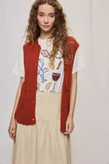 Chaleco de punto color beige, sin mangas, con escote redondo y cierre frontal con botones carey. Presenta diseño calado y silueta amplia.