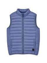 Chaleco acolchado tipo puffer, color azul, con cierre frontal completo, cuello alto y bolsillos laterales. Presenta un pequeño parche decorativo en la parte inferior.