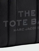 Cartera tipo tote de cuero rojo de la marca Marc Jacobs, modelo The Leather Small Tote Bag. Presenta doble asa superior, correa ajustable y extraíble para el hombro, y el texto grabado en relieve "THE TOTE BAG MARC JACOBS" en el frente.