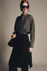 Blusa color gris con finas rayas verticales negras, cuello redondo con lazo del mismo tejido, manga larga con puño, bolsillos de parche en el pecho, bajo asimétrico y cierre frontal oculto con botones.