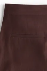 Pantalón sastrero de tiro alto color marrón, modelo tobillero de piernas amplias, con cierre frontal, pretina prolongada con botón y pliegues en la parte delantera y trasera.