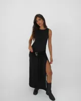 Vestido largo negro sin mangas, con cuello redondo y tajo lateral profundo. Presenta un cinturón incorporado con doble hebilla metálica y flecos largos decorativos en la cintura.