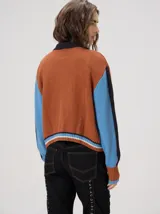 Campera estilo bomber Mutma Satifo, de diseño colorblock con cuerpo en color óxido y mangas celestes con detalles en azul marino. Cuenta con cuello camisero, cierre frontal, puños y ruedo elásticos rayados, y un parche gráfico en el pecho.