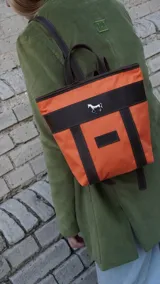 Mini mochila Sierra Mora de diseño estructurado, confeccionada en tela naranja con detalles y correas en cuero marrón. Presenta un aplique metálico plateado con forma de caballo en la parte superior y un parche de cuero con el logo de la marca en el frente.
