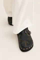 Zueco de cuero con diseño artesanal, presenta una tira con hebilla metálica decorativa, bordados sutiles y múltiples remaches metálicos en la parte frontal. El interior está forrado en cuero.