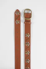 Cinturón de cuero sintético color marrón tostado, con diseño decorativo de flores formadas por tachas metálicas plateadas y doradas, y hebilla metálica en tono bronce envejecido.