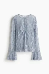 Blusa de encaje color celeste pálido, con cuello redondo y abertura de cerradura con botón forrado. Presenta frunces en el busto y mangas largas con puños anchos de volado.