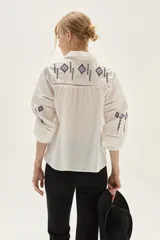 Blusa blanca de algodón con bordados geométricos en tonos negros y lilas, cuello clásico, cierre frontal con botones y mangas largas abullonadas con puños. Presenta detalles de vainillas y calados en el frente y mangas.