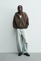 Sudadera con capucha y cierre de cremallera frontal, color marrón oscuro, de corte holgado (boxy fit) y manga larga. Presenta bolsillos frontales de vivo y acabados en rib en puños y bajo.