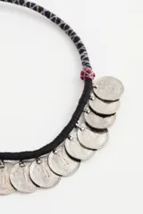 Collar corto de estilo étnico con cordón negro de poliéster y múltiples dijes colgantes en forma de monedas de metal plateado. Presenta detalles de cuentas rojas decorativas a los lados.